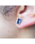 Emerald cut deep blue sapphire stud earring in 14K white gold worn on an ear.