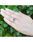 Hand wearing a yellow gold ring with a blue natural sapphire against a green leafy background