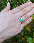Hand wearing a ring with an emerald stone with matching wedding band against a green leafy background.