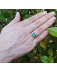 Hand wearing a ring with an emerald stone with matching wedding band against a green leafy background.