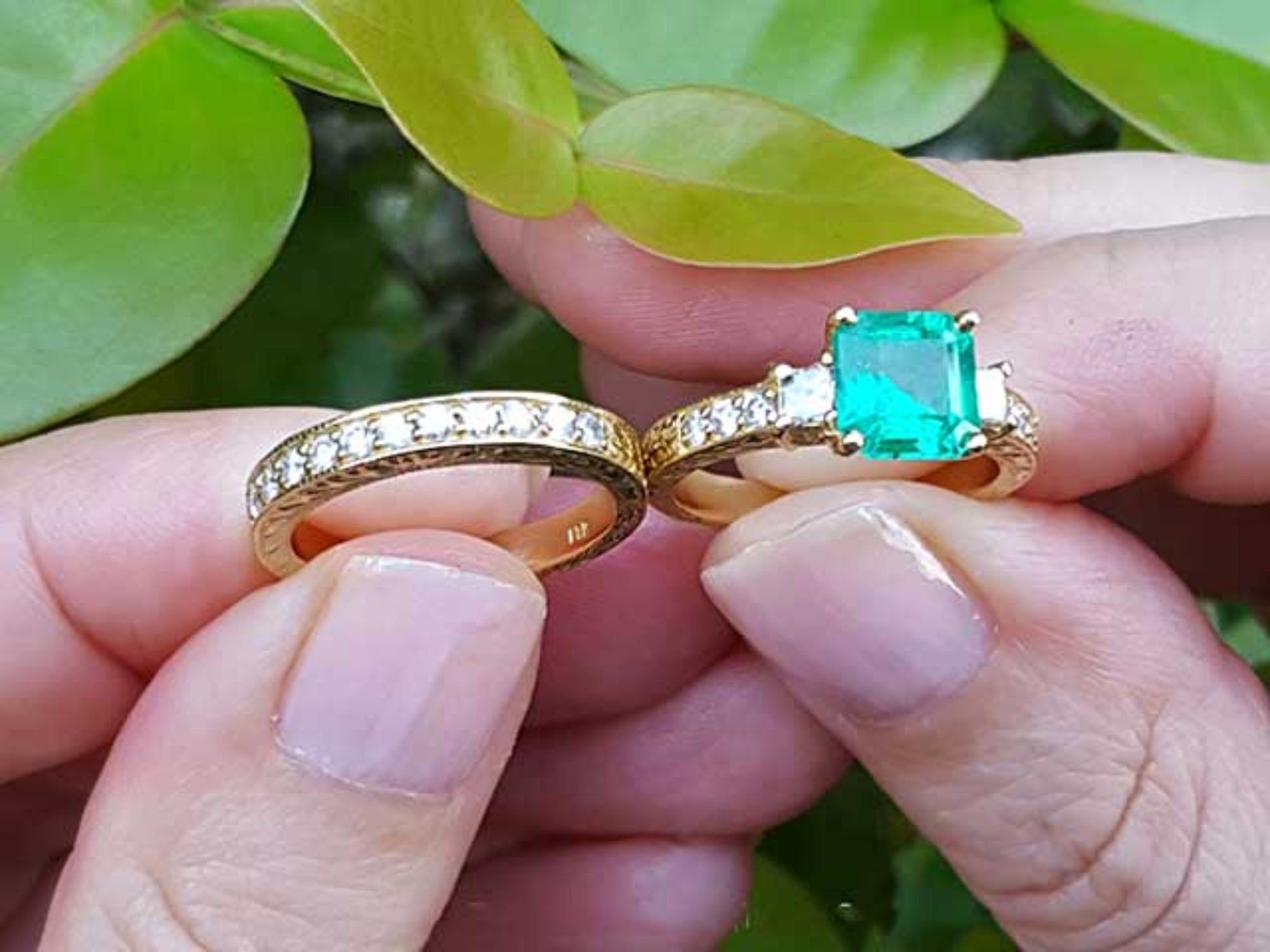Both, emerald ring and matching wedding band held between fingers against a green leafy background.