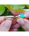 Both, emerald ring and matching wedding band held between fingers against a green leafy background.