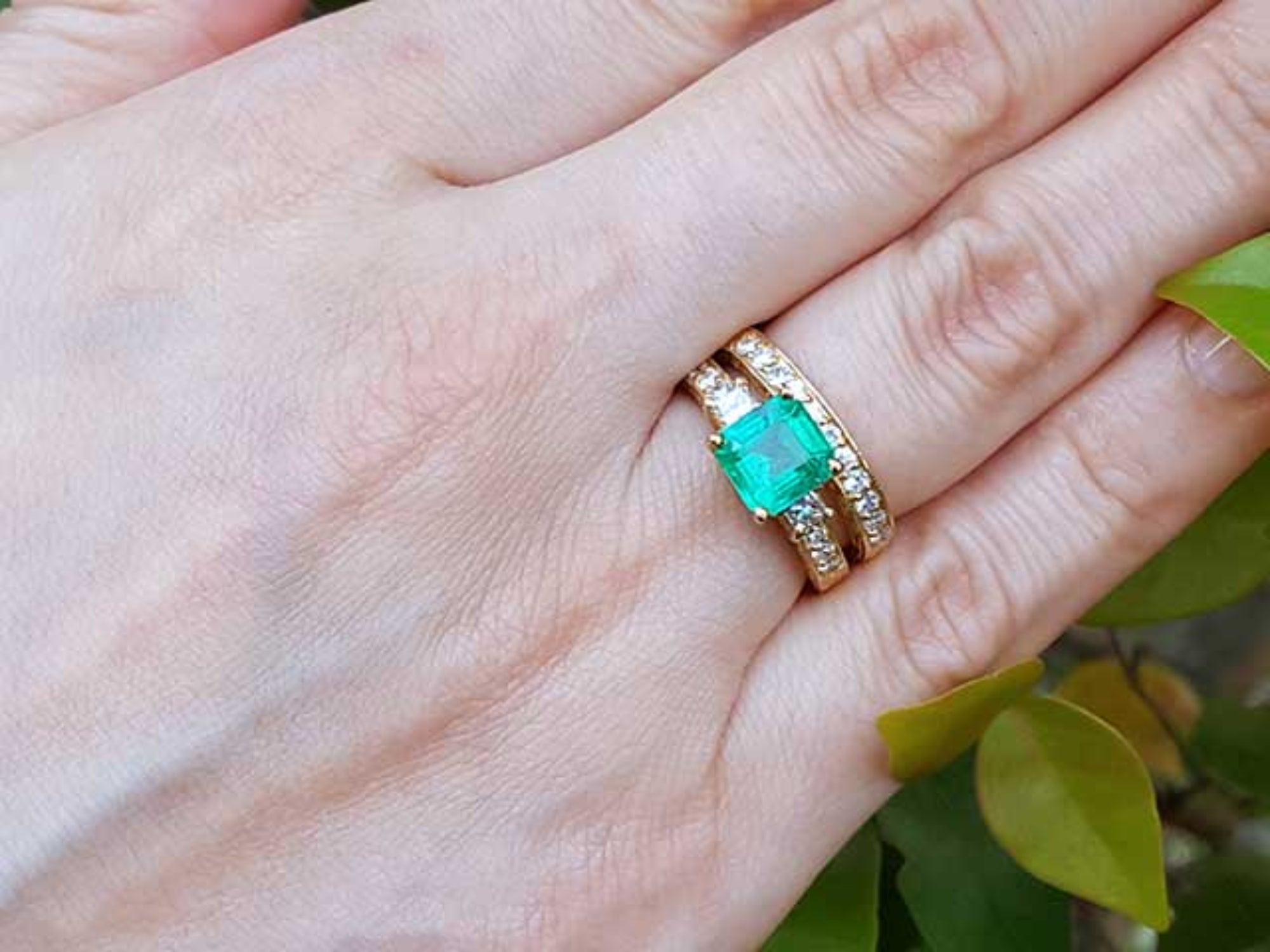 The top view of a yellow gold emerald ring with matching wedding band on a hand, with leaves in the background.