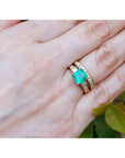 The top view of a yellow gold emerald ring with matching wedding band on a hand, with leaves in the background.