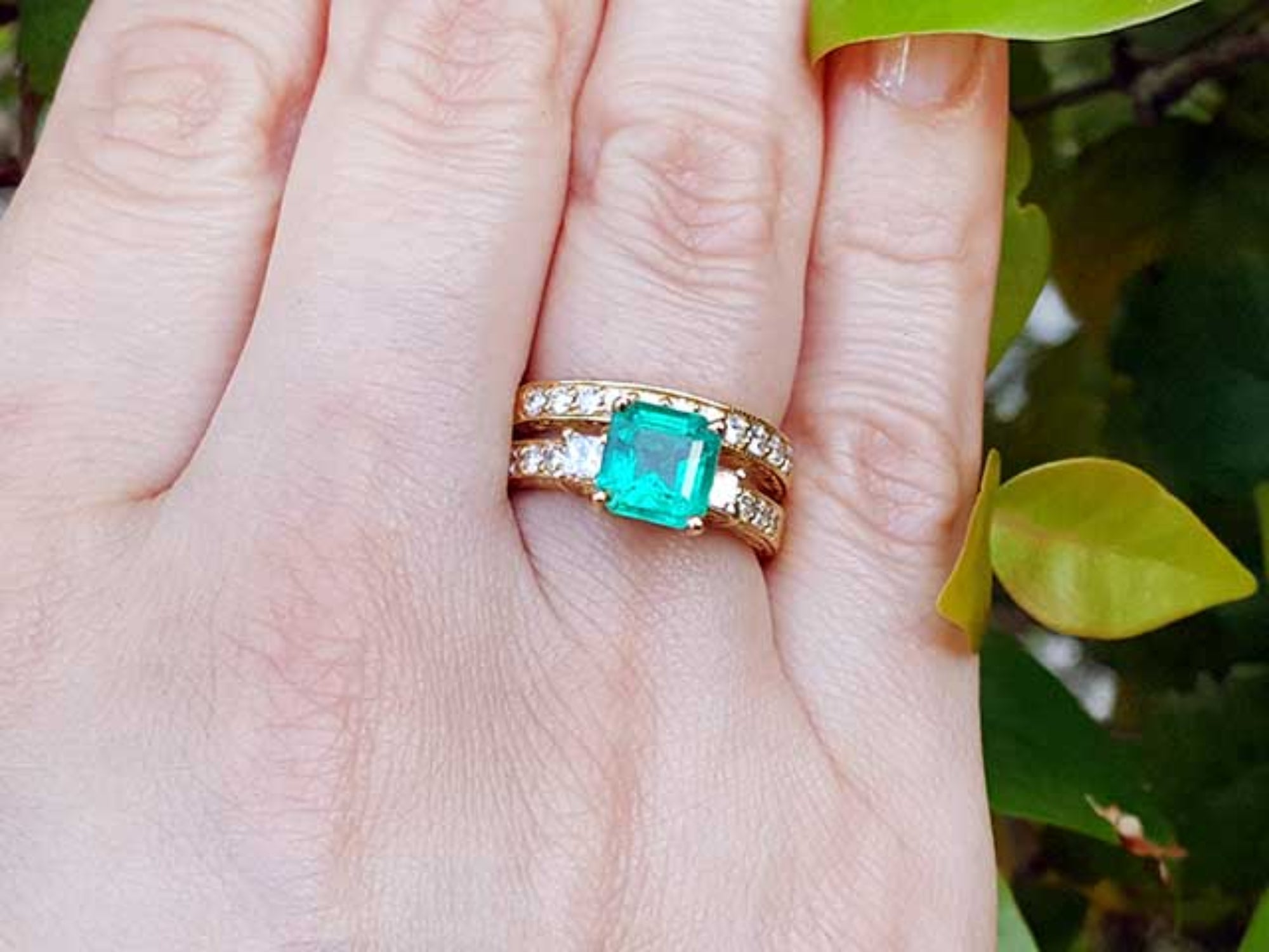A yellow gold emerald ring with the matching wedding band on a hand against a natural background.