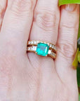 A yellow gold emerald ring with the matching wedding band on a hand against a natural background.