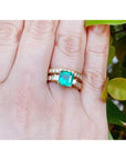 A yellow gold emerald ring with the matching wedding band on a hand against a natural background.