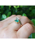 A 18k gold you and me or toi et moi emerald and diamond ring, worn on a person's finger against a background of green foliage.