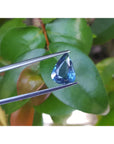 Teardrop-shaped blue natural sapphire held by tweezers against a green leafy background