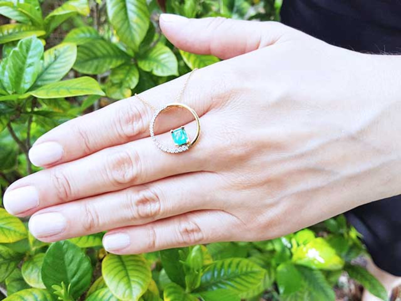 Hand showing an emerald circle of life pendant necklace against a green leafy background.