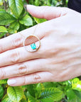 Hand showing an emerald circle of life pendant necklace against a green leafy background.