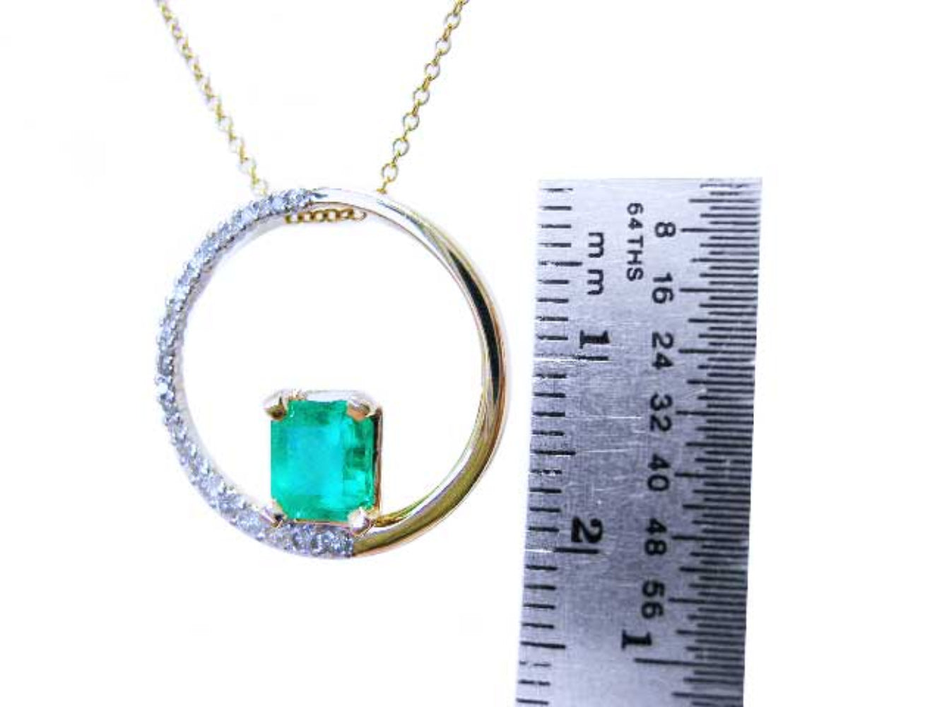 Gold circle of life pendant with a square natural emerald and diamonds, next to a ruler for scale.