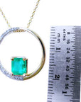 Gold circle of life pendant with a square natural emerald and diamonds, next to a ruler for scale.