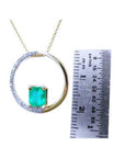 Gold circle of life pendant with a square natural emerald and diamonds, next to a ruler for scale.
