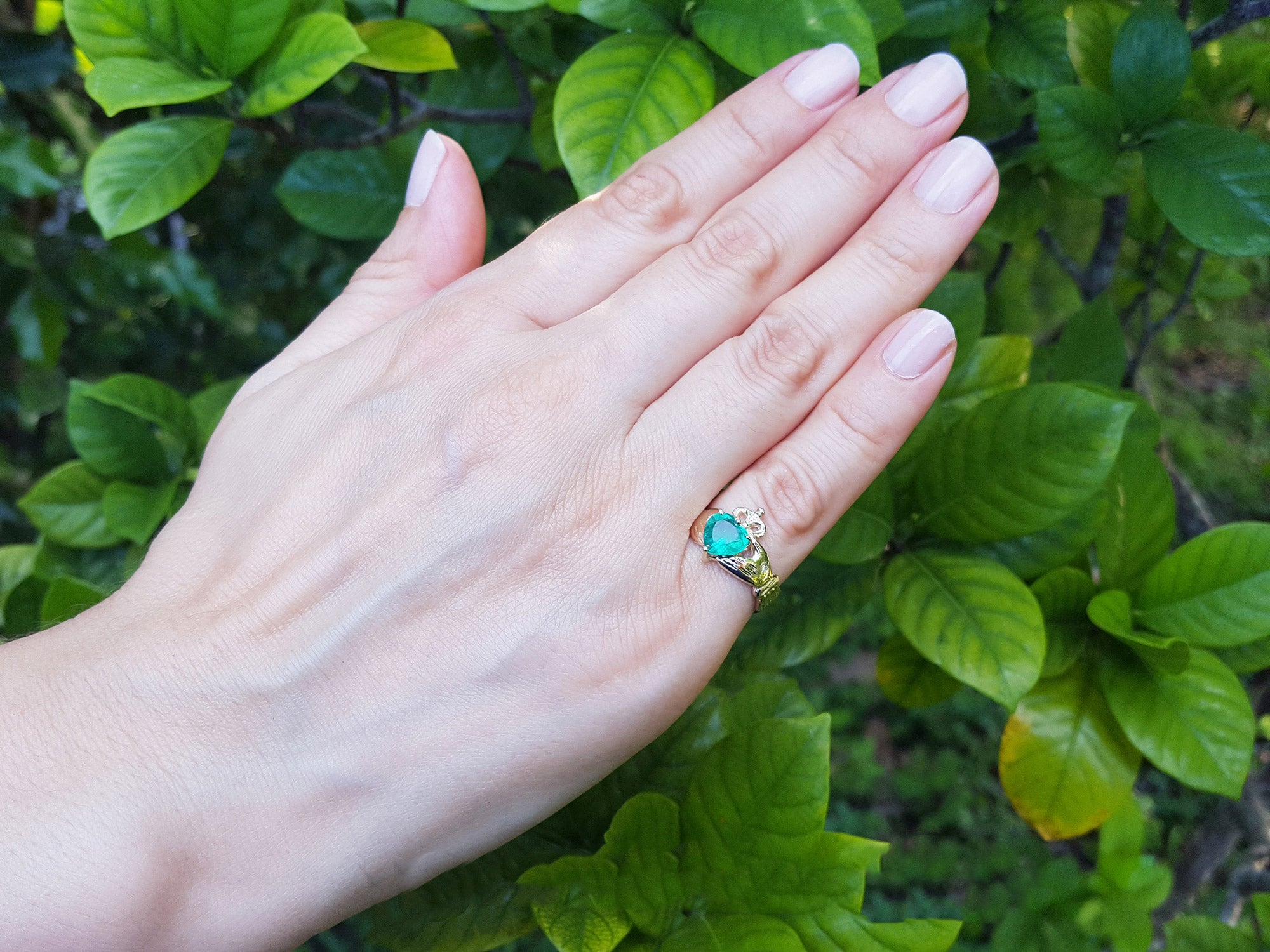 Hand wearing a heart emerald claddagh ring against a natural leafy background.