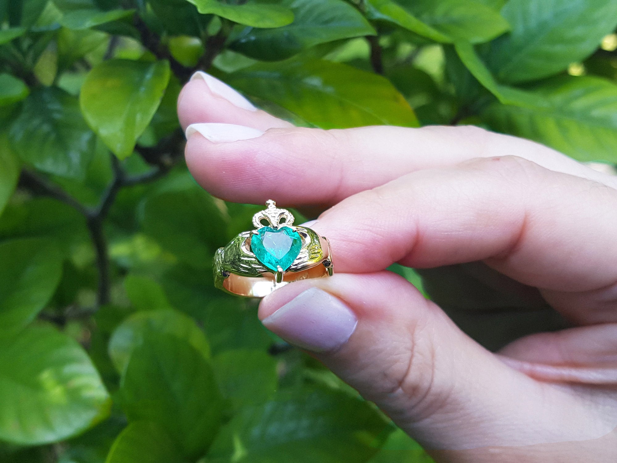 A heart emerald claddagh ring being held between fingers on a green leafy background.