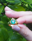 A heart emerald claddagh ring being held between fingers on a green leafy background.