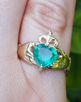 A yellow gold heart emerald claddagh ring on a finger against a blurry green background.