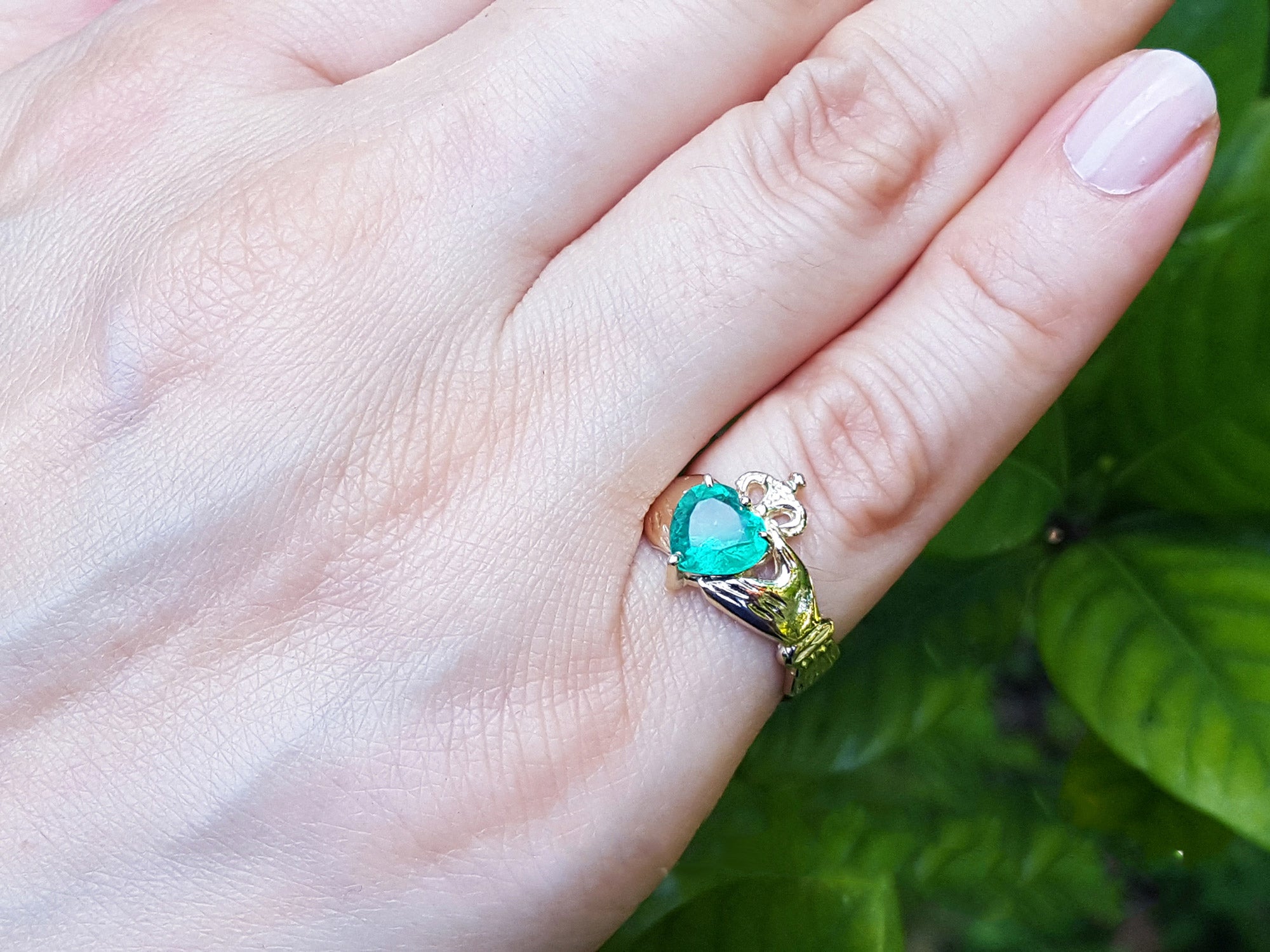 A heart cut emerald claddagh ring being worn on a woman's hand on a leafy background.