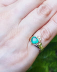 A heart cut emerald claddagh ring being worn on a woman's hand on a leafy background.