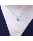A close sight of a gold necklace with a oval Muzo emerald gemstone pendant on a person wearing a dark blue top.