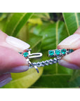 Close-up of hands holding a white gold tennis bracelet with round cut emeralds against a leafy background.