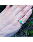 Hand wearing a 18k yellow gold ring with a square colombian emerald and trillion diamonds against a natural background.