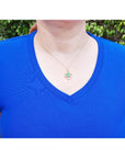 14K gold clover pendant with a square emerald and diamonds, worn on a woman in a blue shirt.