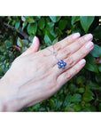 A woman's hand displaying a cluster of natural sapphires pendant white gold necklace on a leafy background