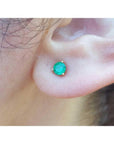 Medium deep green emerald stud earring in 14K yellow gold with three prongs, worn on an ear.