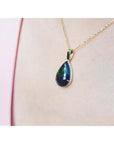 Teardrop-shaped Wello black opal pendant on a gold chain against a pink background