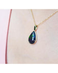Teardrop-shaped Wello black opal pendant on a gold chain against a pink background