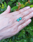 Hand displaying a pair of Muzo mines emerald stud earrings against a green leafy background