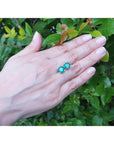 Hand displaying a pair of Muzo mines emerald stud earrings against a green leafy background