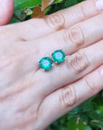Natural round cut emerald stud earrings on a hand with a green leafy background