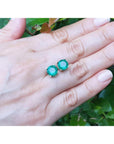 Natural round cut emerald stud earrings on a hand with a green leafy background