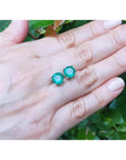 Round bluish-green Colombian emerald stud earrings in 14K yellow gold on a hand.