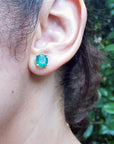 Close-up of an ear wearing a Colombian emerald stud earring with a blurred green leafy background