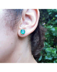 Close-up of an ear wearing a Colombian emerald stud earring with a blurred green leafy background