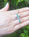 Hand holding a white gold cross necklace with natural emeralds against a green leafy background.