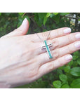 Hand holding a white gold cross necklace with natural emeralds against a green leafy background.