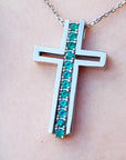 A white gold cross pendant with round cut natural emeralds on a chain, worn on a person's neck.