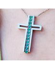 A white gold cross pendant with round cut natural emeralds on a chain, worn on a person's neck.