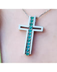 A white gold cross pendant with round cut natural emeralds on a chain, worn on a person's neck.