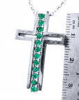 A white gold cross pendant with natural emeralds next to a ruler for scale on a white background.