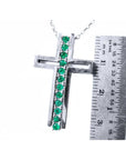A white gold cross pendant with natural emeralds next to a ruler for scale on a white background.