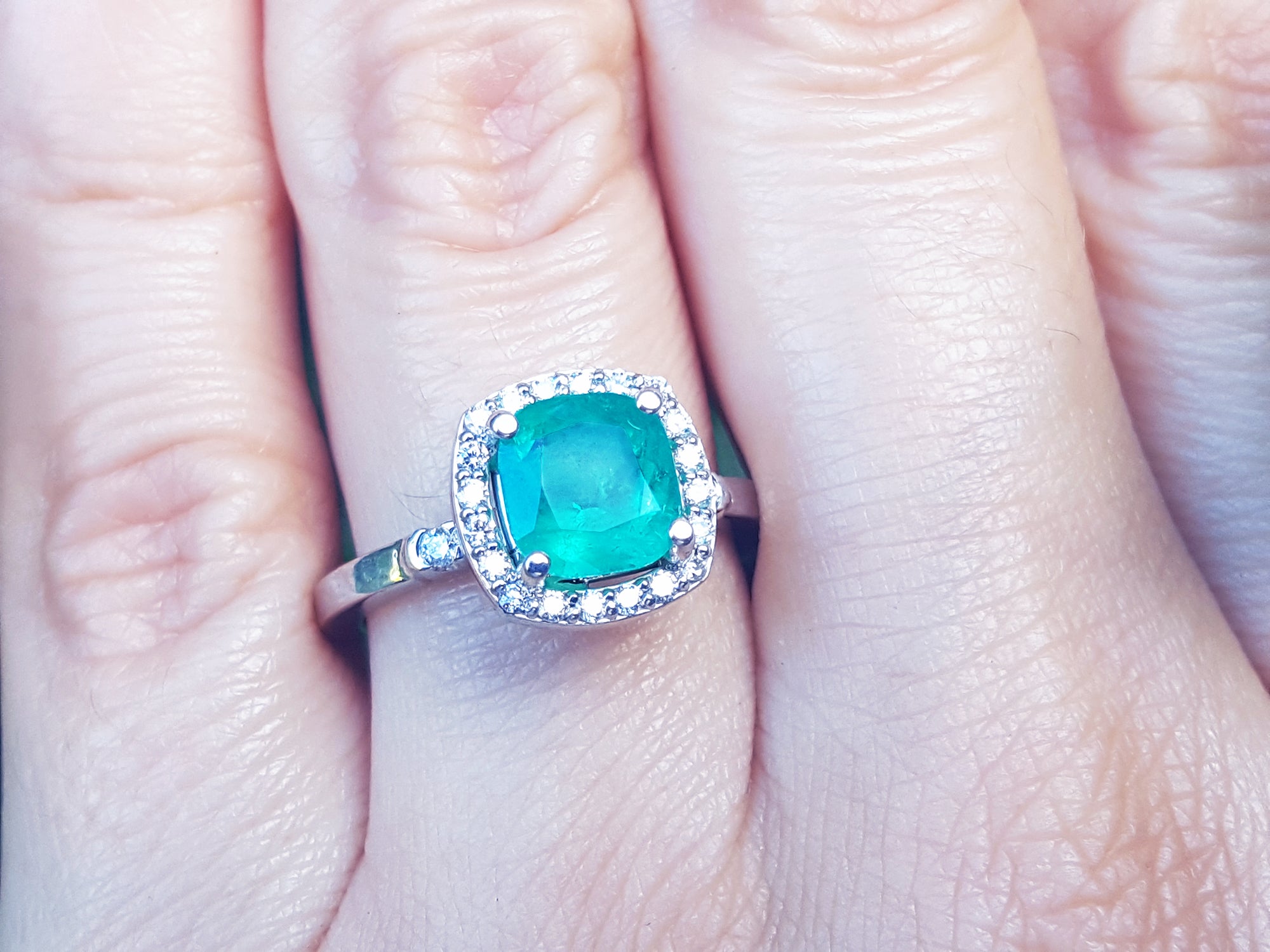 A close-up of a cushion cut emerald surrounded by a halo diamond in white gold on a person's hand.