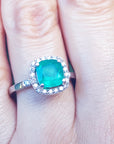 A close-up of a cushion cut emerald surrounded by a halo diamond in white gold on a person's hand.