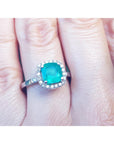 A close-up of a cushion cut emerald surrounded by a halo diamond in white gold on a person's hand.