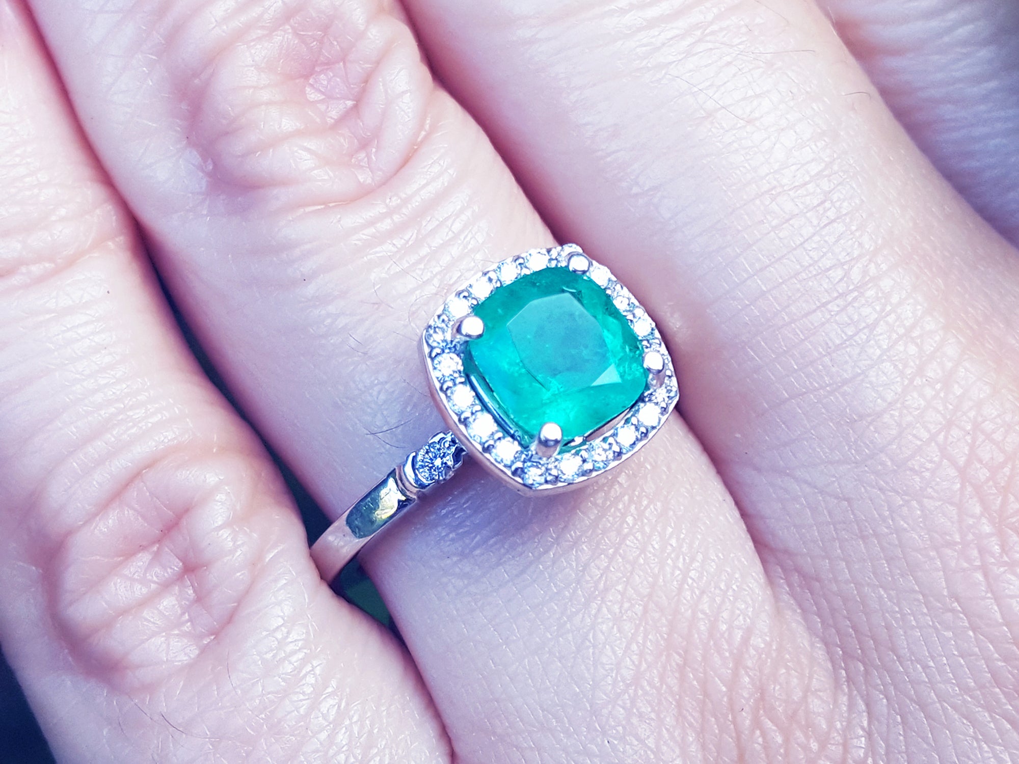 A natural Colombian emerald cushion cut with a halo diamond in white gold on a finger.